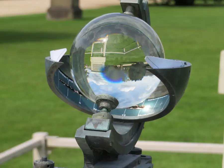 A glass sphere sundial stands on a metal frame, reflecting an upside-down image of the green lawn, sky, and nearby structures in its center.