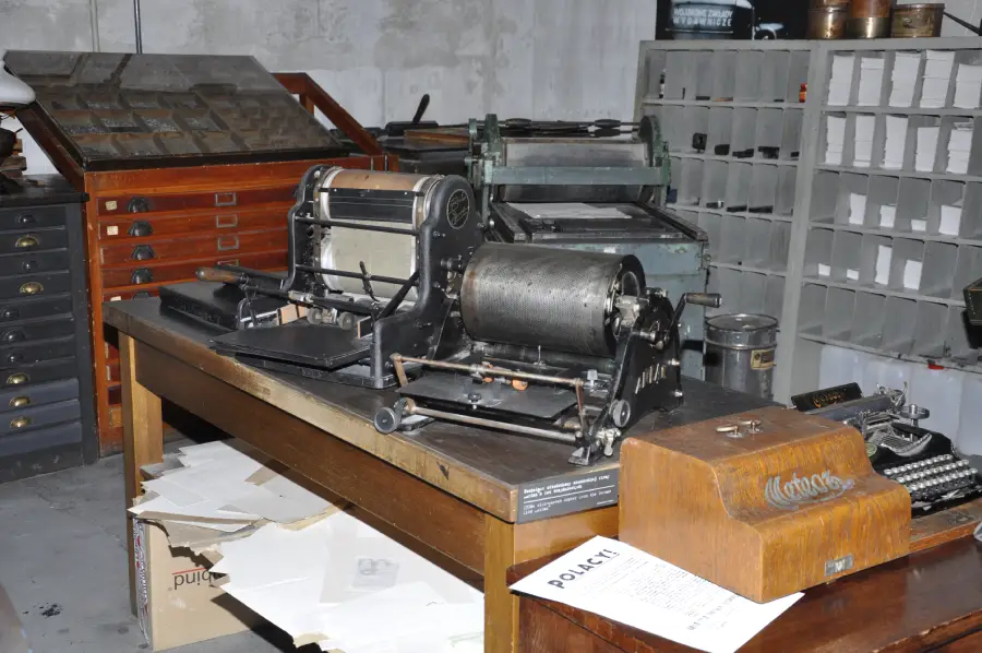 A vintage duplicating machine sits on a wooden table in an old office, surrounded by papers, filing drawers, and shelves with compartments.