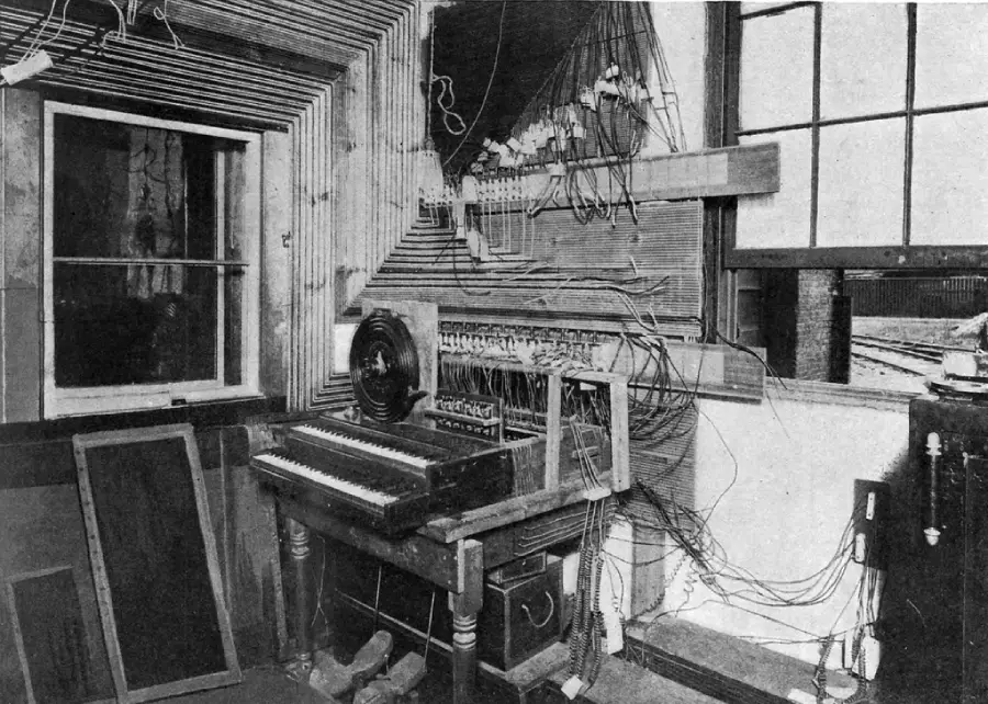 A vintage room with a keyboard device, many wires, and a control panel mounted on the wall, next to large windows.