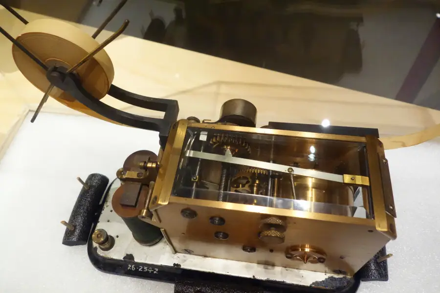 A close-up view of an antique mechanical telegraph machine with exposed gears and a paper tape reel, displayed on a white surface.