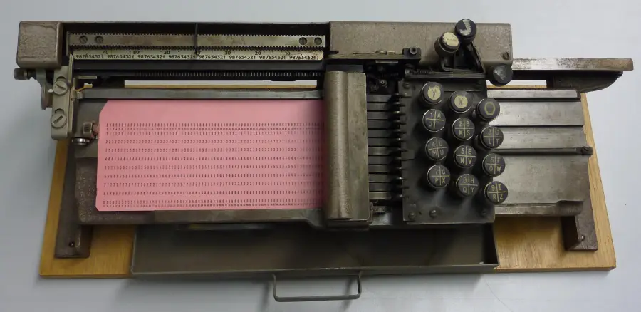 An old punched card machine with round keys, a pink punched card inserted, and metal levers, mounted on a wooden base.