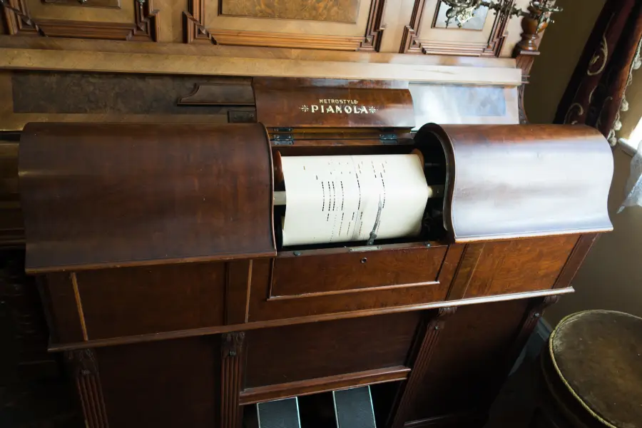 A close-up of a wooden player piano with a visible paper roll inserted, featuring perforated holes for automatic music playback. The top cover is partially open, revealing the mechanism inside.
