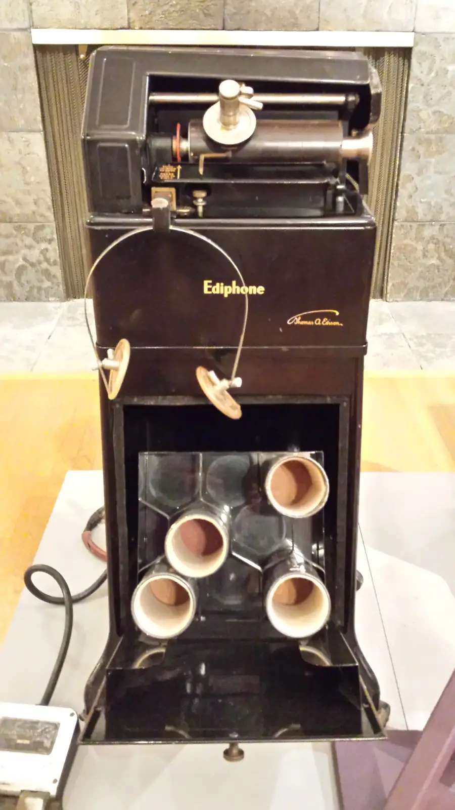 An antique black Edison Ediphone dictation machine with four empty cylinder holders and attached earphones, displayed indoors on a wooden floor.