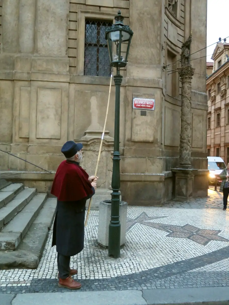 An older man in a hat and cape uses a long pole to light a streetlamp beside a historic stone building with steps and mosaic pavement.