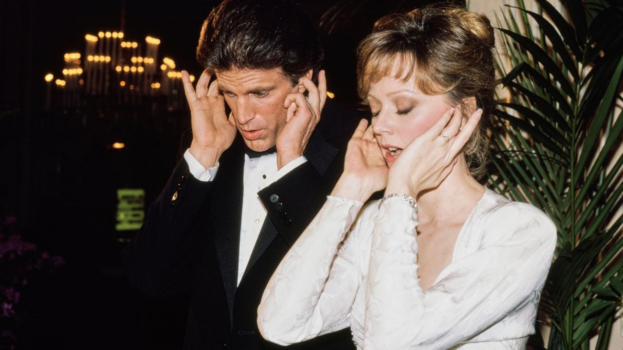 A man in a tuxedo and a woman in a white dress cover their ears with their hands, standing closely together, possibly at a formal event with soft lighting and plants in the background.