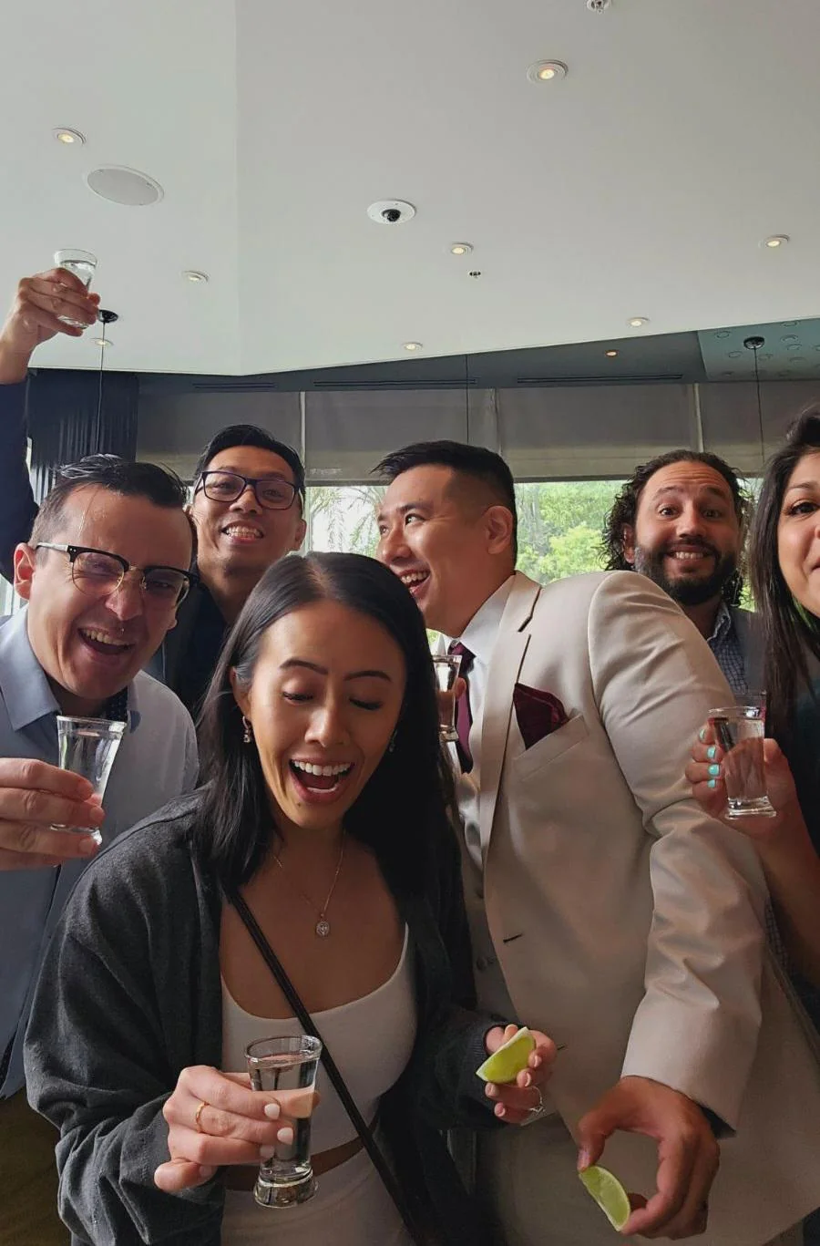 A group of people are smiling and laughing indoors, holding shot glasses and lime wedges, celebrating together. One person is dressed in a white suit.