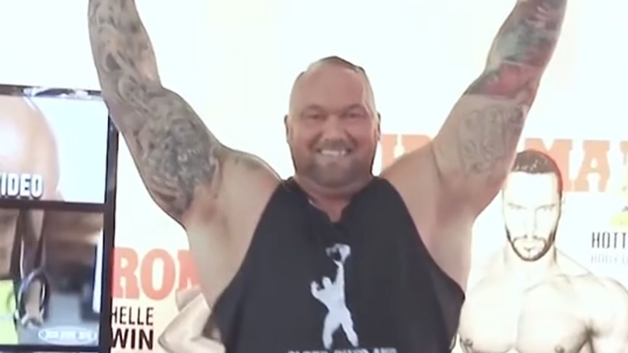 A muscular man with tattooed arms raises both arms and smiles while wearing a sleeveless black shirt, standing in front of a background with fitness posters.