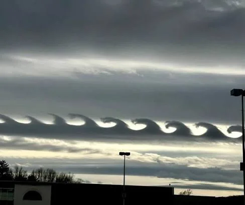 Wave-shaped clouds form a unique pattern in the sky above a cityscape with silhouetted buildings and streetlights, under an overcast atmosphere.