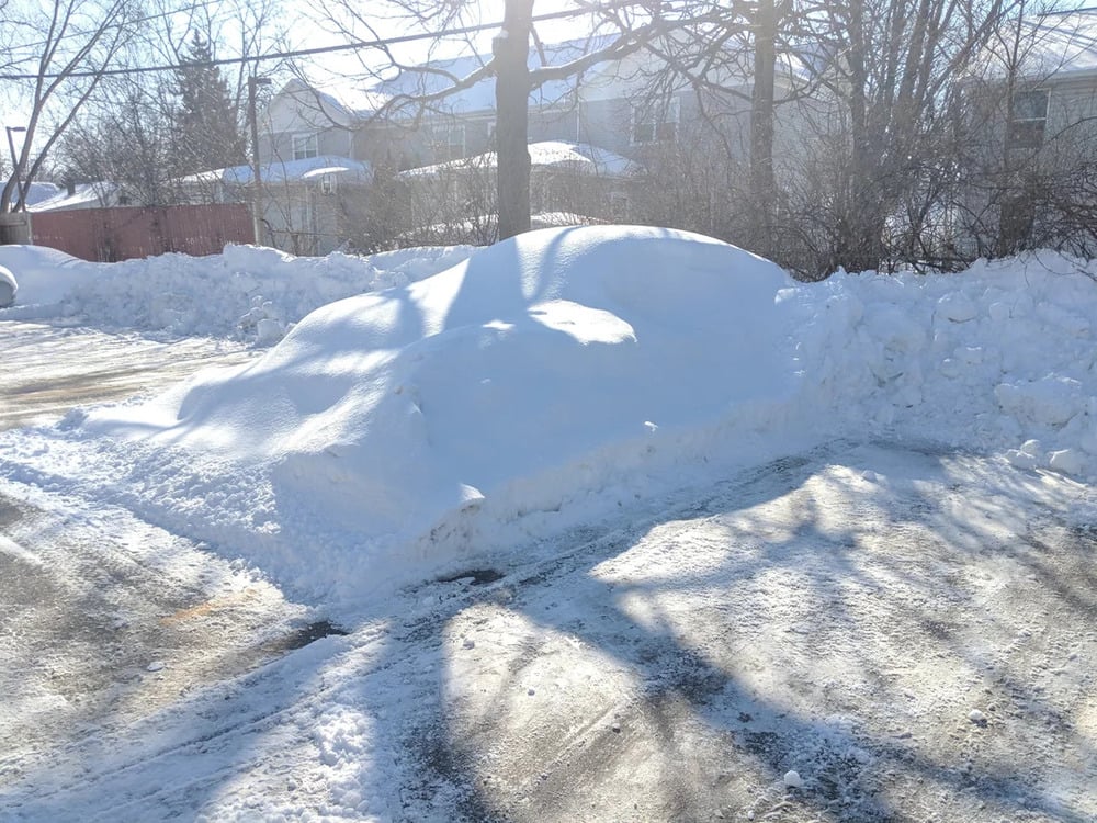 A car is completely covered by a thick layer of snow in a parking lot, surrounded by snowbanks and bare trees, with sunlight casting shadows on the ground.