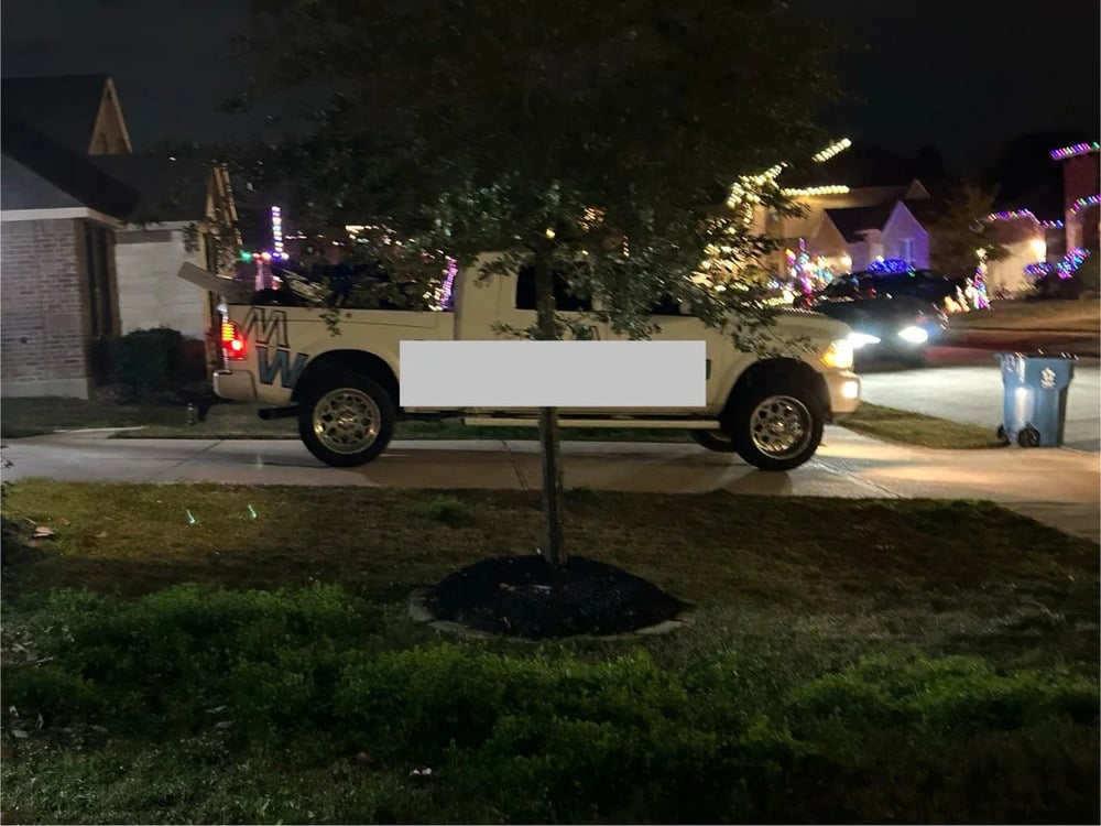 A white pickup truck is parked in a driveway at night, partially blocked by a small tree. The street and nearby houses are lit with colorful holiday lights. A blue recycling bin is visible on the curb.