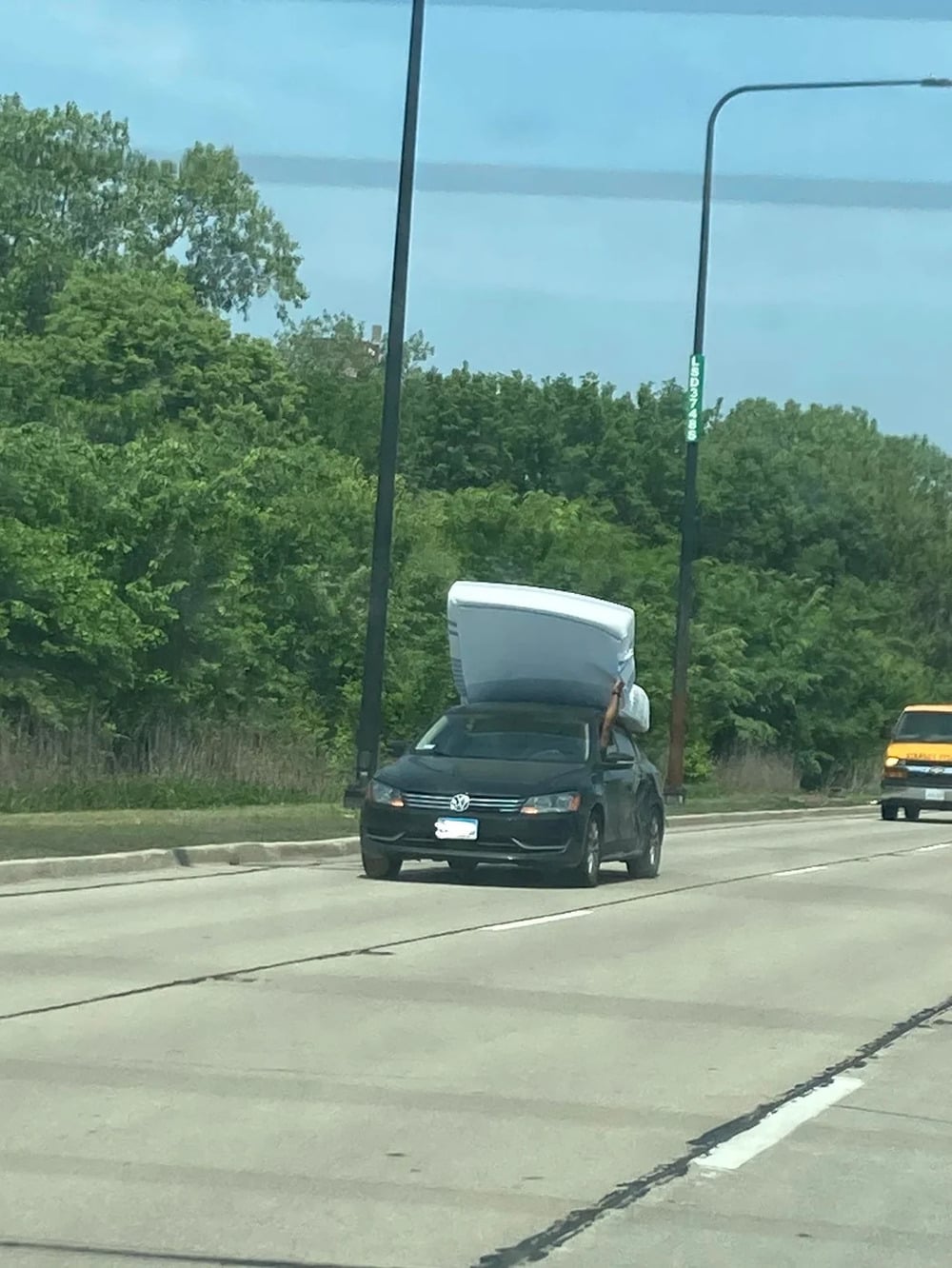 A person drives a car with a mattress strapped to the roof, holding it with one hand out the window while driving on a road lined with trees.