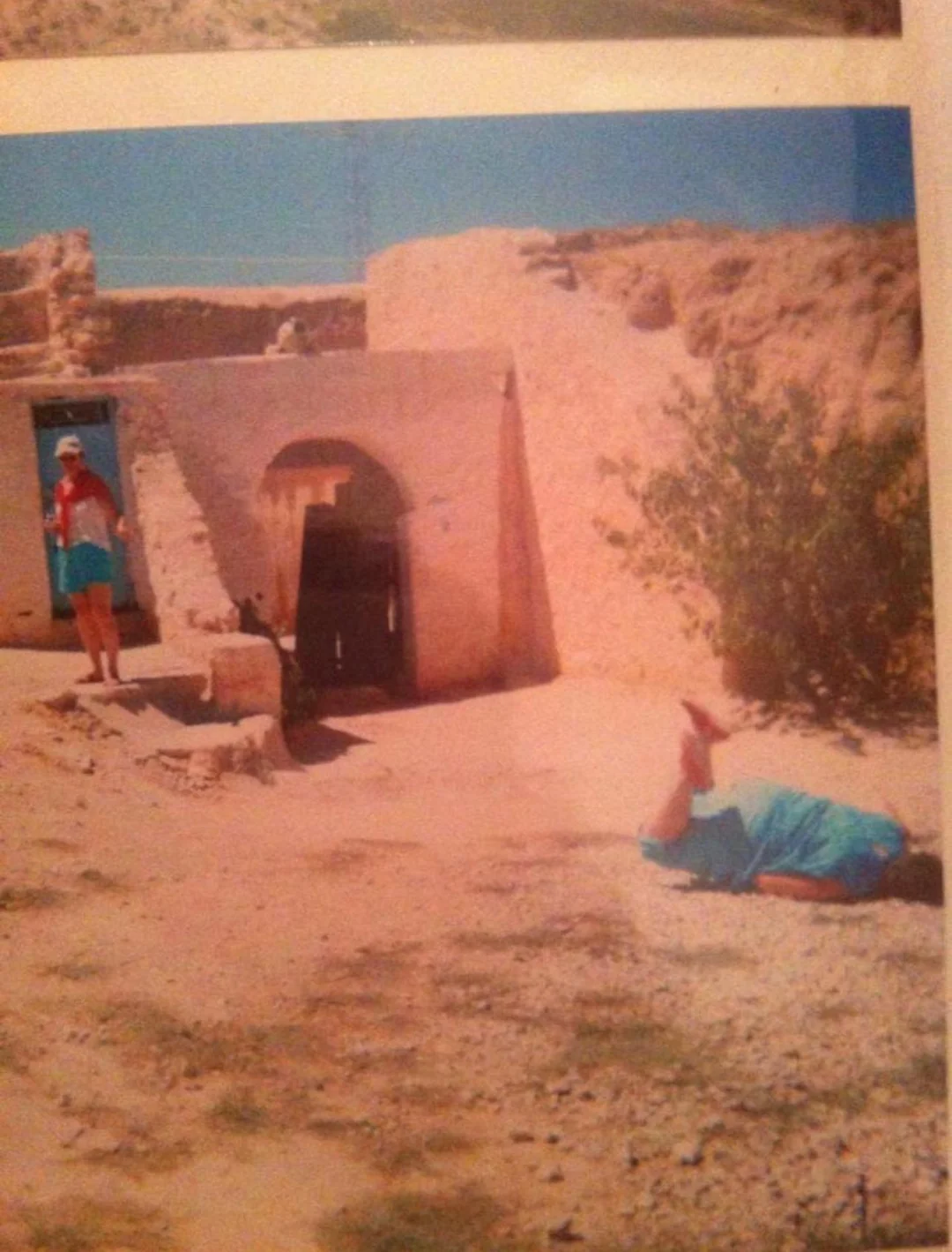 Two people in blue outfits are outside a rustic stone building in a desert setting; one stands near a doorway, and the other lies on the ground, appearing to have fallen. The sky is clear and bright.