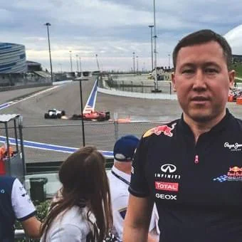 A man in a racing team shirt stands at a motorsport event, with race cars speeding on a track and spectators nearby. The sky is cloudy and stadium structures are visible in the background.