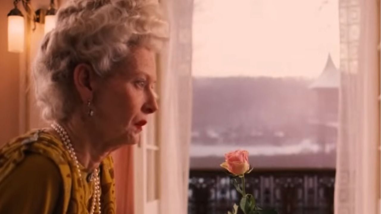 Elderly woman with curly white hair and pearl necklace gazes at a single pink rose in a vase by a window, with a soft outdoor scene in the background.