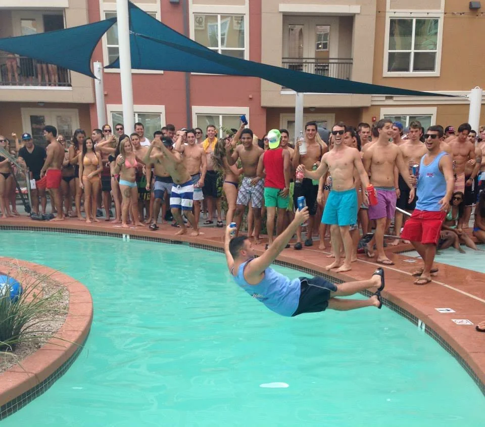 A man in a blue tank top falls backward into a pool while a large crowd of people in swimwear watch and cheer at a pool party.