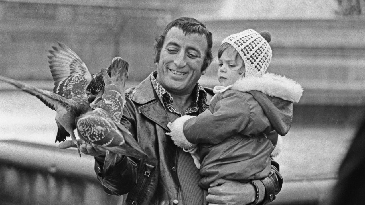 A man in a leather jacket holds a young child and smiles while feeding pigeons. The child, bundled in a coat and knit hat, looks at the birds. The background features steps and stone structures.