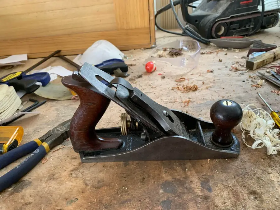 A metal hand plane rests on a messy workbench with wood shavings, pliers, and other tools scattered around.