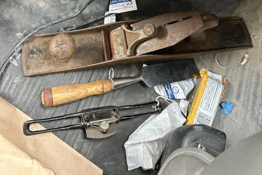 A wood plane, chisel, and scraper are on a dusty floor alongside a snack bar wrapper, napkin, and miscellaneous items.