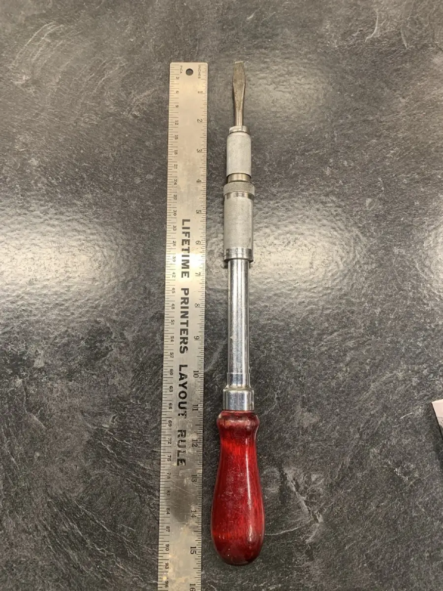 A metal ruler next to a hand tool with a red wooden handle and a flat blade, likely a screwdriver or chisel, on a dark surface. The tool is about 13 inches long.