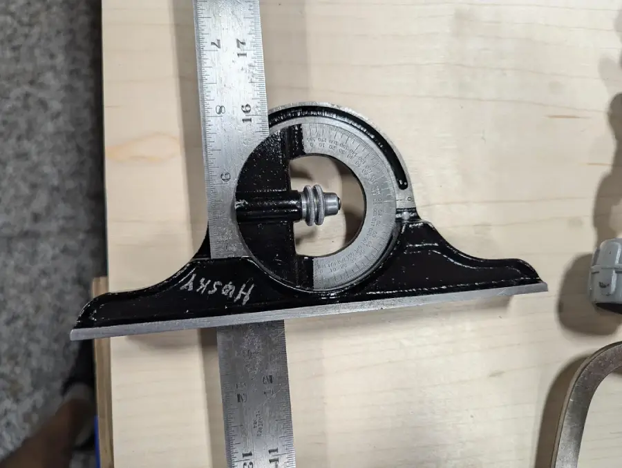 A metal combination square with a black base and ruler is placed on a light wood surface, showing measurements and markings.