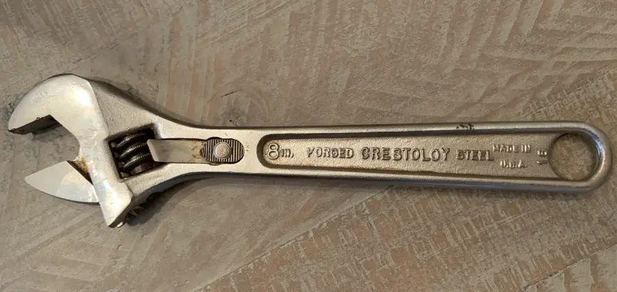 A silver adjustable wrench with “Forged Crestoloy Steel” inscribed on the handle, lying on a wooden surface.