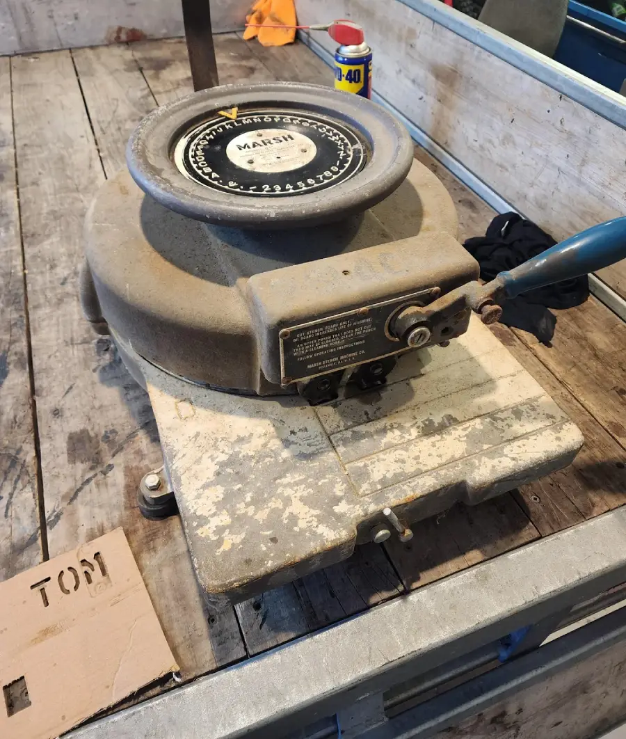 A vintage MARSH label machine sits on a worn wooden workbench, with a can of WD-40 and various objects nearby. The machine shows signs of age and use.