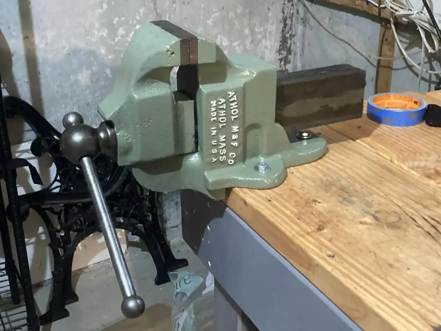 A metal bench vise mounted on the edge of a wooden workbench in a workshop with tools and cables nearby.