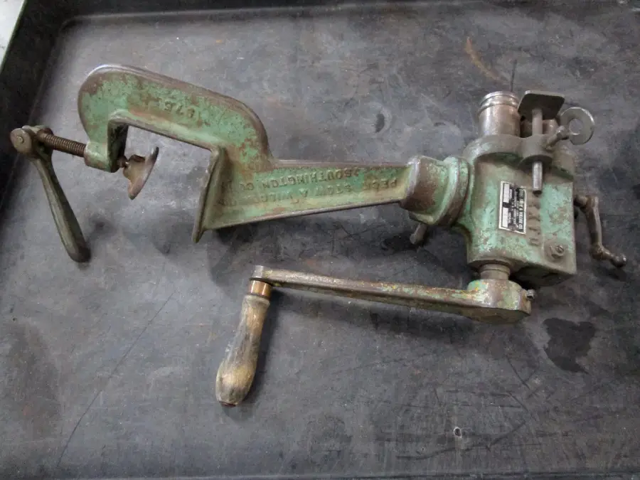 A vintage green metal handheld tool with a clamp on one end and a rotating handle on the other, lying on a dark surface. The tool has some rust and wear, indicating frequent use.