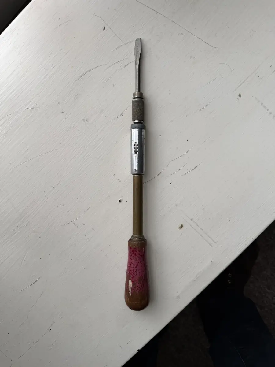 A vintage hand tool with a flathead screwdriver tip, a metal shaft, and a worn wooden handle, lying on a white surface.