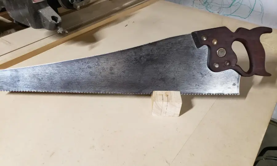 A hand saw with a dark wooden handle rests on a light-colored work surface, its blade placed on top of a small rectangular wooden block.