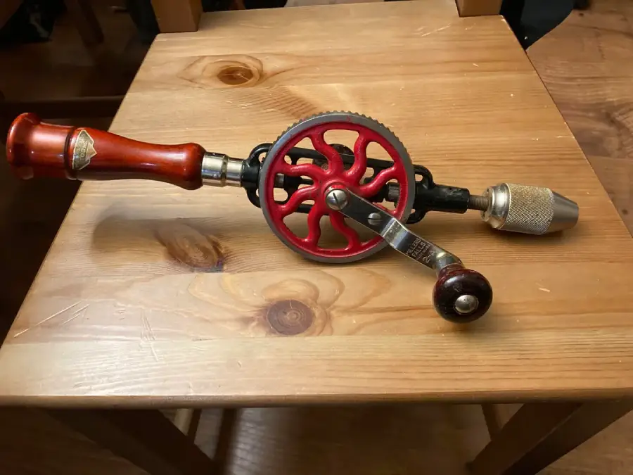 A vintage hand-crank drill with a red gear and polished wood handle rests on a wooden table.