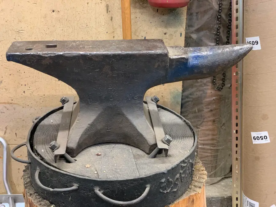 A heavy metal anvil sits securely strapped onto a circular base, placed on a cut tree stump in a workshop. Various tools and materials are visible in the background.