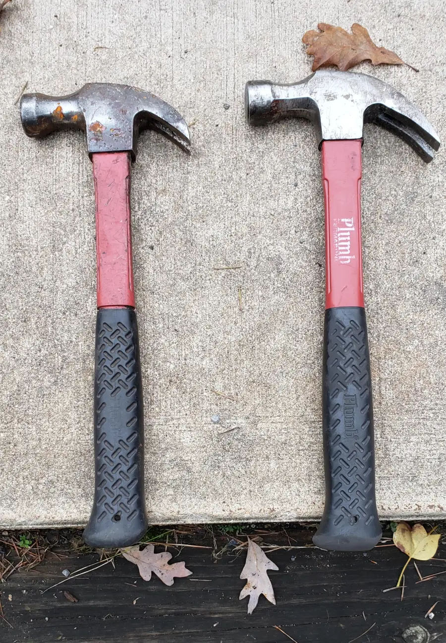 Two claw hammers with red handles and black grips lying side by side on a concrete surface, surrounded by a few fallen leaves.