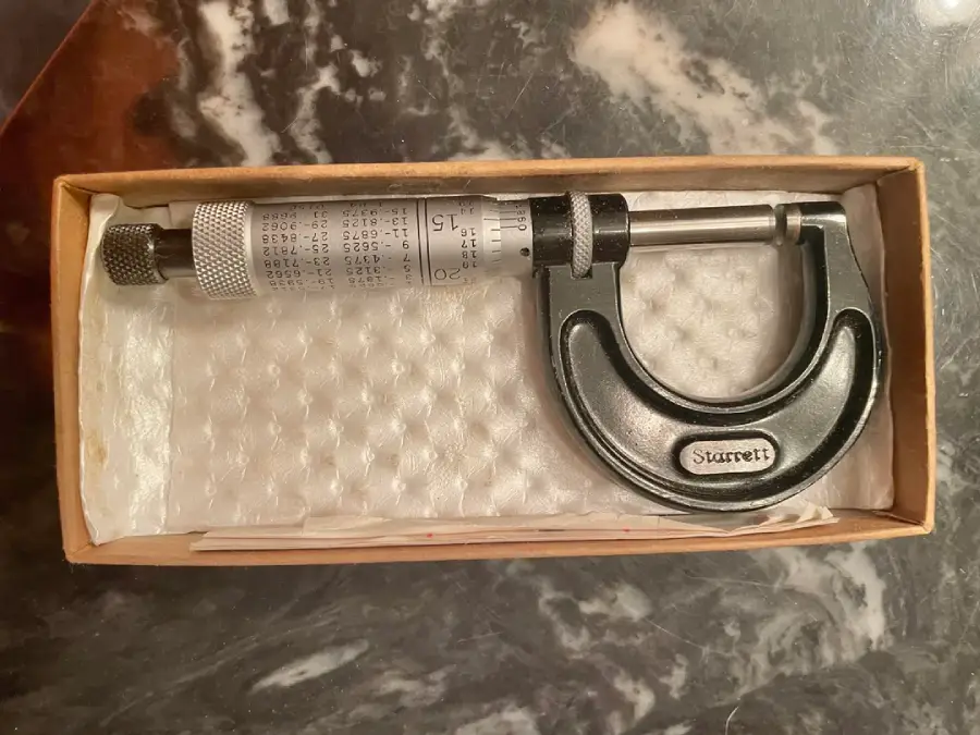 A metal micrometer in a box lined with white padding, placed on a dark, marbled surface. The micrometer is labeled “Starrett” and is used for precise measurement.