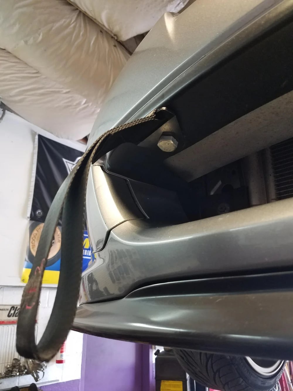 A close-up of a car’s front bumper with a fabric tow strap attached, hanging down. The car is in a garage with a banner and tools visible in the background.