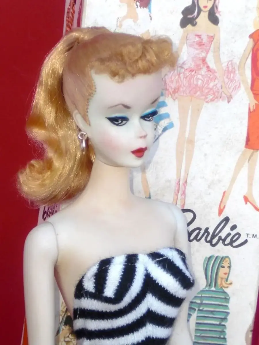 Vintage Barbie doll with blonde hair in a ponytail, blue eye shadow, red lips, and a black-and-white striped dress, posed in front of Barbie-themed packaging with illustrations.