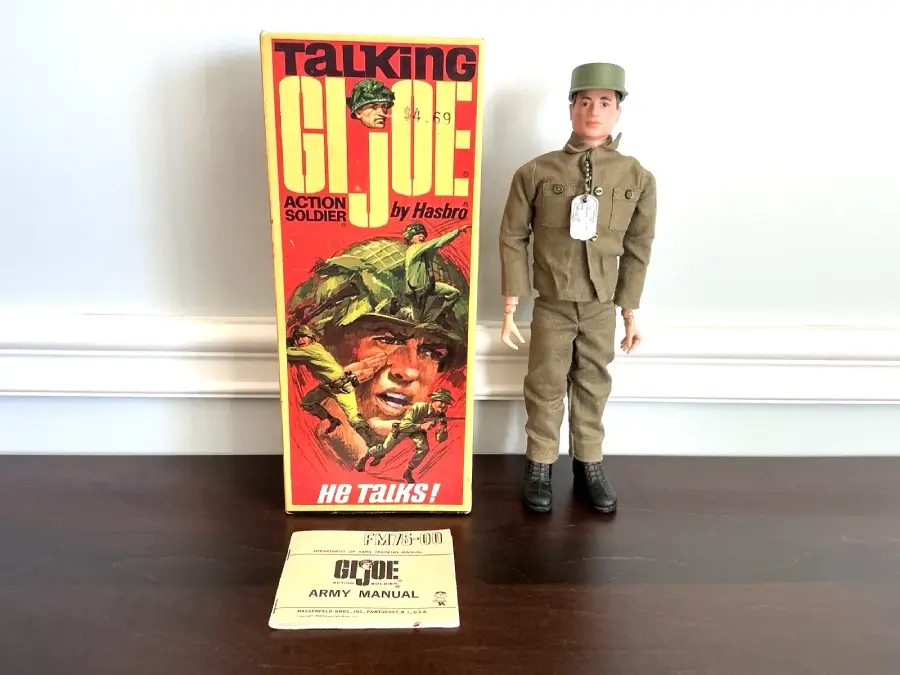 A vintage Talking GI Joe action figure stands next to its colorful box and an army manual on a dark table against a white wall.