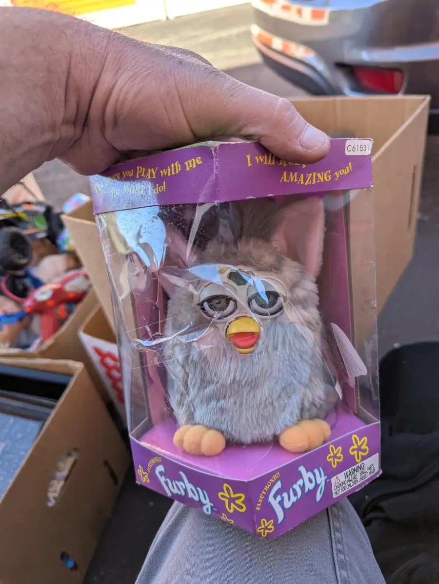 A hand holds a boxed gray Furby toy with its plastic packaging cracked. The box is purple with yellow text and sits among other items in a cardboard box outdoors.