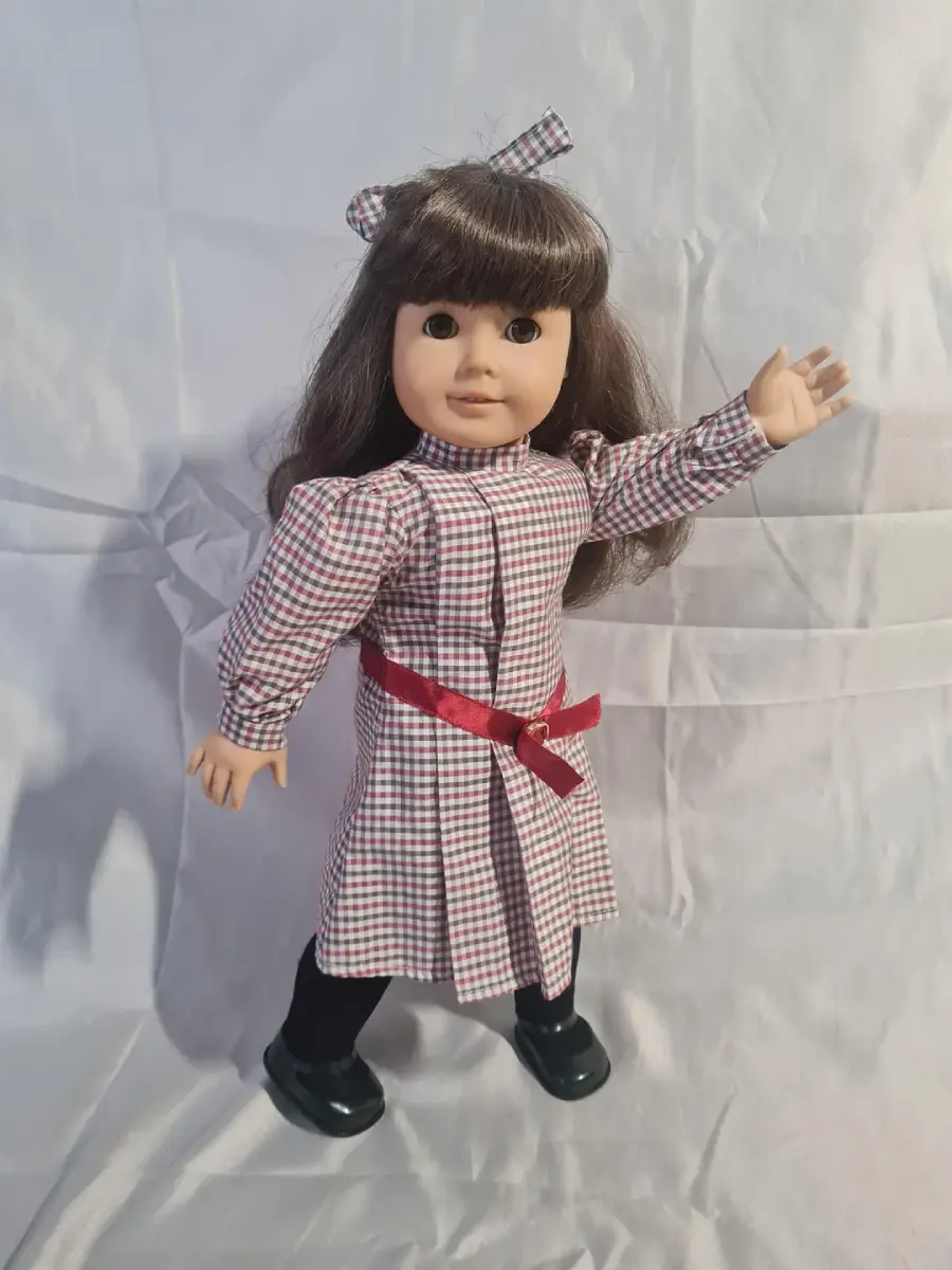A doll with long brown hair, wearing a red and white checkered dress with a red belt, black shoes, and a matching hair bow, stands on a white fabric background, raising one arm.