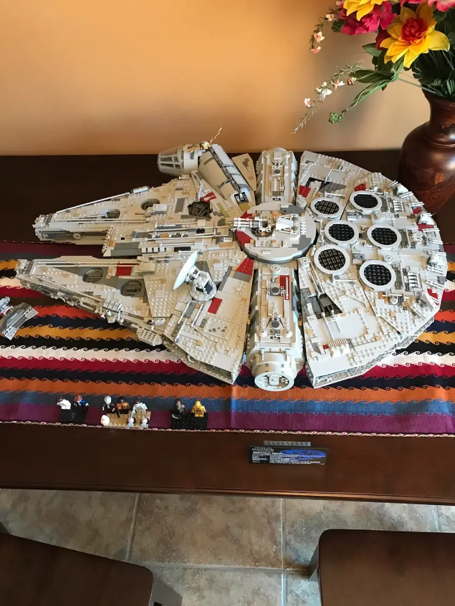 Large LEGO Millennium Falcon model on a striped table runner, with mini-figures displayed in front and a vase of flowers in the background on a wooden table.