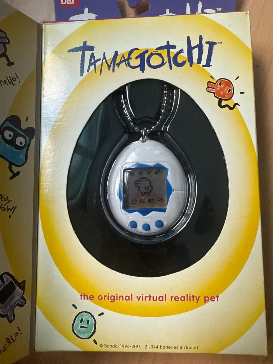 A packaged Tamagotchi virtual pet on a yellow and white background, featuring cartoon icons and the text “the original virtual reality pet” at the bottom. The device has a keychain and a black and white screen.