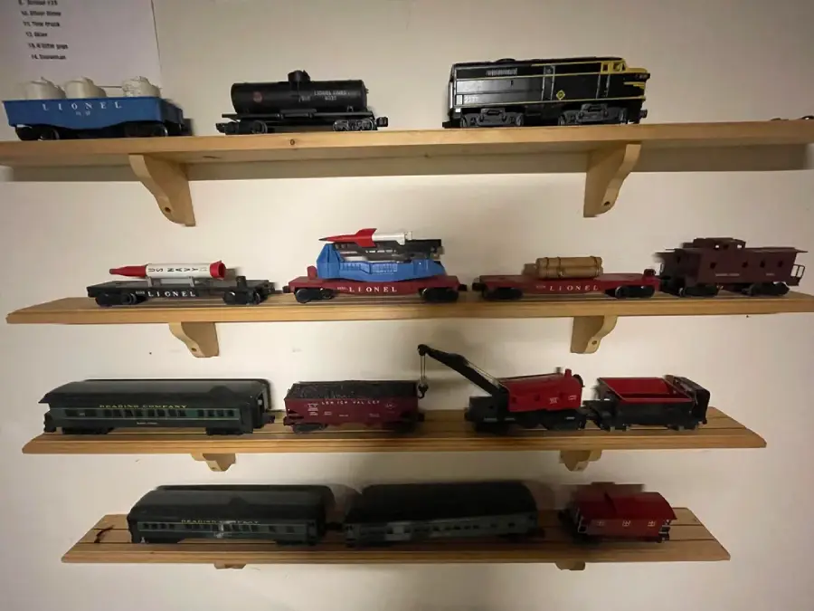 Four wooden shelves display a collection of vintage model trains, including engines, cars, and cabooses, arranged neatly against a beige wall under warm lighting.