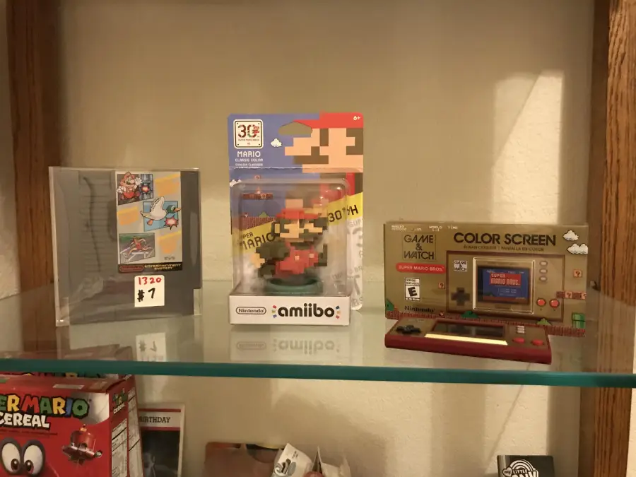 A shelf displays a Mario Amiibo in original packaging, a boxed Super Mario Bros. Game & Watch Color Screen, and a Nintendo game manual with price tags, all neatly arranged on a glass shelf.