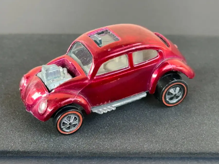 A small red toy car resembling a Volkswagen Beetle with exposed engine parts, large rear wheels, and a sunroof, displayed on a dark surface.