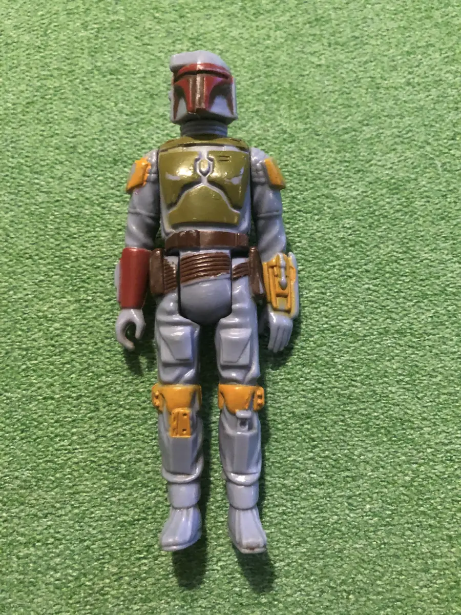 An action figure in gray and green armor with a helmet, brown belt, and yellow details lies on a textured green surface.
