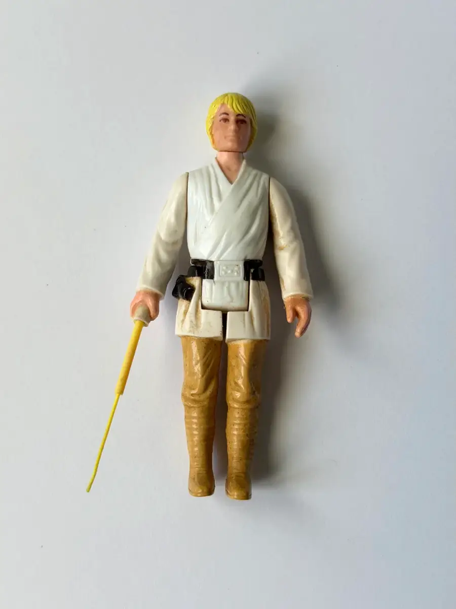 Action figure with blond hair, white tunic, tan pants, and brown boots holding a yellow lightsaber on a plain white background.