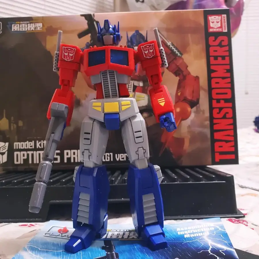 A red, blue, and gray Optimus Prime action figure stands in front of its Transformers model kit box with artwork of the character and bold “Transformers” text on the right side.