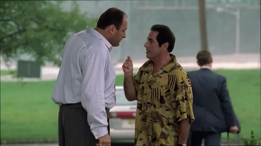Two men stand outside facing each other, one in a white shirt and the other in a patterned shirt, engaged in a tense conversation. A third man with a suitcase walks away in the background.