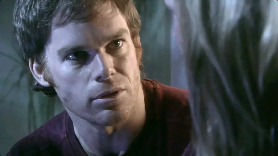 A man with a serious expression looks intently at someone out of frame. The lighting is dim, casting blue tones on his face. He wears a maroon shirt and has short brown hair.