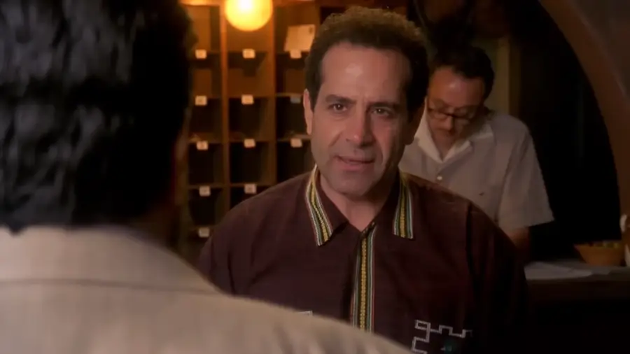 A man wearing a dark patterned shirt stands indoors, facing another person whose back is visible. In the background, a person in glasses writes at a desk near cubbyholes and a lit bulb.